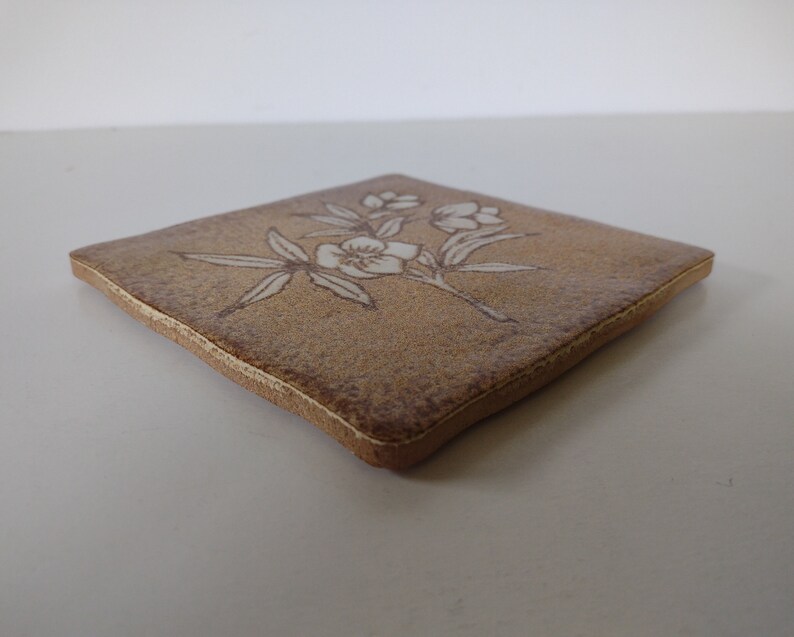 May include: A square, tan ceramic coaster with a floral design. The flowers and leaves are white, and the coaster has a slightly textured surface. The edges are a darker brown.