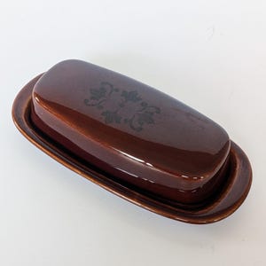 May include: A brown ceramic butter dish with a lid. The lid has a floral design. The dish is oval-shaped and has a glossy finish. The butter dish is a kitchenware item.