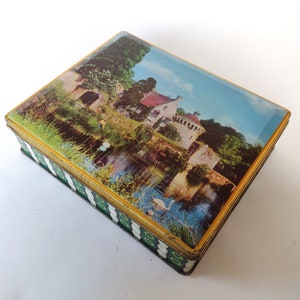 Puede incluir: Una caja de hojalata vintage con una imagen colorida de un edificio de piedra con un foso y un cisne en el agua. La caja tiene un borde a rayas verde y blanco con detalles dorados.