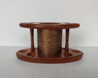 May include: A wooden pipe rack with a round base and a central cylinder for storing pipes. The rack has six individual slots for pipes and is made of dark brown wood.