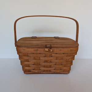 May include: A brown woven picnic basket with a wooden lid and handle. The basket has a rectangular shape and a wooden latch. The handle curves over the top, and the lid is secured with two small wooden pegs.