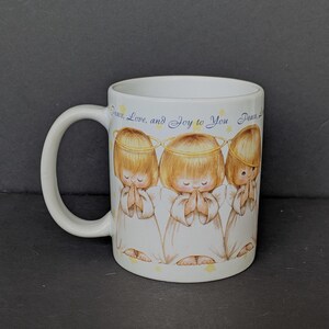 May include: White ceramic mug featuring three cartoon angels with halos, hands clasped in prayer. The angels have blonde hair and wear white robes. The mug has the text "Peace, Love, and Joy to You" in a script font.