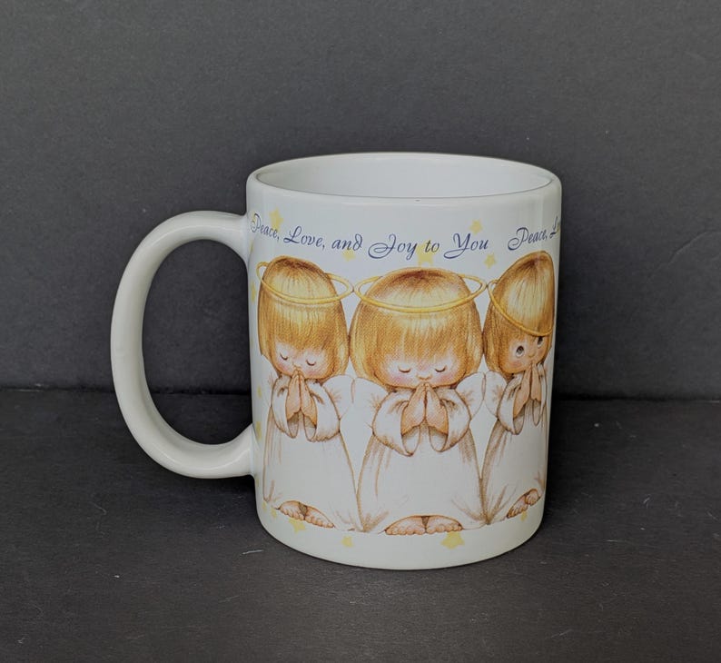 May include: White ceramic mug featuring three cartoon angels with halos, hands clasped in prayer. The angels have blonde hair and wear white robes. The mug has the text "Peace, Love, and Joy to You" in a script font.