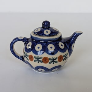 May include: A small ceramic teapot featuring a cream-colored base with a hand-painted pattern of dark blue circles, orange dots, and green leaf-like motifs. The teapot includes a matching lid with a knob handle and a curved spout on the side.