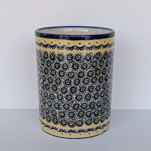 Może przedstawiać: Cylindryczny ceramiczny pojemnik na przybory z niebieskim wnętrzem i żółto-niebieskim wzorem spiralnym. Pojemnik ma żółty pasek u góry i u dołu z niebieską ozdobną obwódką.