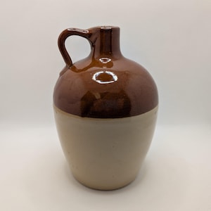 May include: A vintage stoneware jug with a rounded body and a handle. The top portion is a rich brown color, while the bottom half is a creamy beige. This classic piece is likely used for storing liquids and adds a rustic touch to any decor.