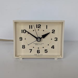 May include: A rectangular Westclox electric alarm clock with a cream-coloured face, black numerals, and black hour and minute hands. The clock features a thin second hand, a clear plastic cover, and the text "WESTCLOX electric" and "MADE IN U.S.A."