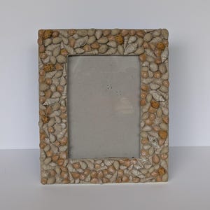 May include: A rectangular photo frame, adorned with a mosaic of seashells in beige, tan, and cream hues. The frame's border is densely packed with shells of various sizes and shapes, creating a textured surface. The central opening is a neutral grey.