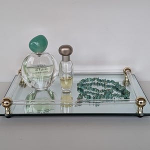 May include: A mirrored vanity tray with gold-colored accents holds a perfume bottle with a teal cap, a small perfume bottle, and a turquoise beaded necklace. The tray has a rectangular shape and clear acrylic handles.