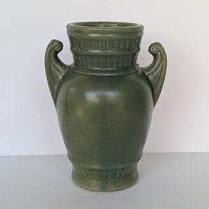 May include: A green ceramic vase with two curved handles. The vase has a wide base and a narrower neck, with decorative ridges near the top and bottom. The overall shape is reminiscent of classical pottery.