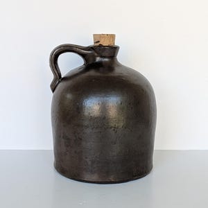 May include: A dark brown ceramic jug with a rounded body, a single side handle, and a light-colored cork stopper inserted into the narrow neck. The surface of the jug has a mottled, uneven texture with subtle variations in the dark brown glaze.