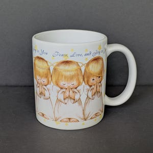 May include: White ceramic mug featuring three cartoon angels with halos, hands clasped in prayer, and wearing white robes. The mug has the text "Joy to You Peace, Love, and Joy to You" in blue script and yellow stars.