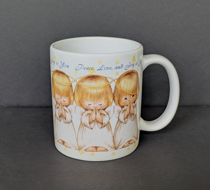 May include: White ceramic mug featuring three cartoon angels with halos, hands clasped in prayer, and wearing white robes. The mug has the text "Joy to You Peace, Love, and Joy to You" in blue script and yellow stars.