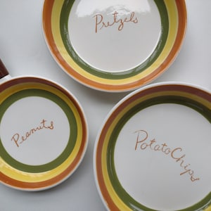 May include: Three ceramic serving dishes with green, yellow, and orange stripes. The dishes are labeled "Pretzels", "Peanuts", and "Potato Chips".