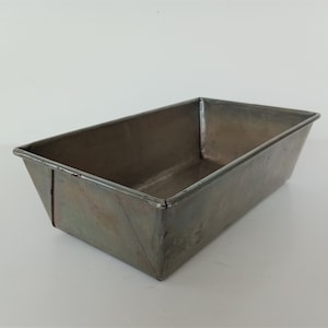 Può includere: Uno stampo per pane vintage in metallo con una finitura grigia leggermente sbiadita. Lo stampo ha una forma rettangolare con lati leggermente inclinati.