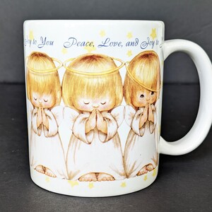 May include: White ceramic mug featuring three cartoon angels with halos, hands clasped in prayer, and wearing white robes. The mug has the text "Joy to You Peace, Love, and Joy to You" in blue script and yellow stars.