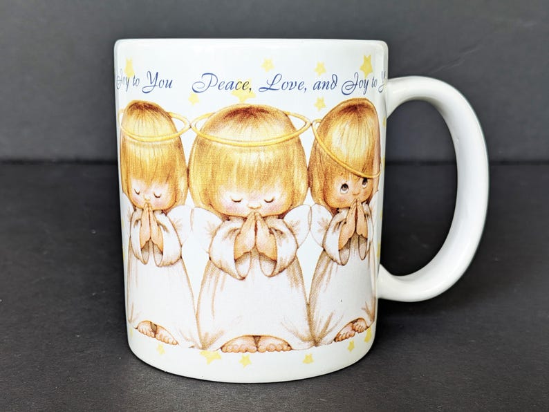 May include: White ceramic mug featuring three cartoon angels with halos, hands clasped in prayer, and wearing white robes. The mug has the text "Joy to You Peace, Love, and Joy to You" in blue script and yellow stars.