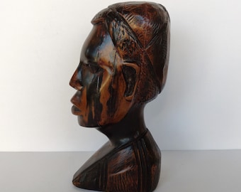 May include: A dark brown wooden sculpture of a person's head and shoulders. The sculpture is carved in a realistic style and has a detailed face. The person is wearing a hat.