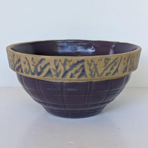 May include: A round ceramic bowl with a deep purple glazed interior and a matching purple exterior featuring a grid pattern. The upper rim is a contrasting tan color with a repeating dark purple leaf-like pattern embossed around the circumference.
