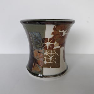 May include: A ceramic vase with a flared shape, featuring a dark brown rim and a base. The vase is decorated with a patchwork design of brown, white, green, and blue floral and geometric patterns. The vase is on a white surface.
