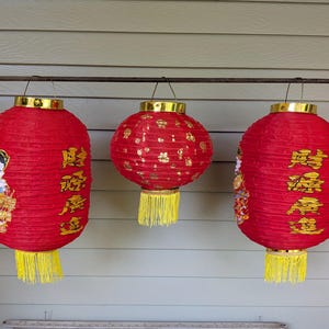 May include: Three red paper lanterns hanging from a horizontal bar. The two outer lanterns are cylindrical with gold tassels, featuring gold Chinese characters and decorative figures. The centre lantern is spherical with gold patterns and a yellow tassel.