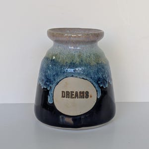 Pode incluir: Um vaso de cerâmica com base larga e gargalo estreito, apresentando um esmalte reativo que transita de um preto escuro na base para um azul profundo e verde claro, com um medalhão circular central cor de creme exibindo a palavra DREAMS.