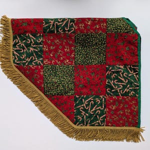 Op de afbeelding: Een kerstboomrok met een patchwork-ontwerp. De rok heeft rode en groene vierkanten met zuurstok-, poinsettia- en andere vakantiepatronen. Het heeft een gouden franjebies rond de rand.