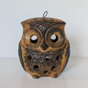 May include: A ceramic owl-shaped lantern featuring a mottled brown and black glaze. The piece has two large circular eye openings and several smaller circular cutouts across the chest area. A thin metal wire loop is attached to the top.