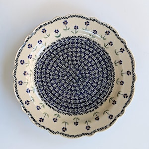 May include: A round ceramic plate with a scalloped edge, featuring a cream-colored background. The center is decorated with a dense pattern of small white flowers on a dark blue field, while the rim displays a border of individual blue flowers.
