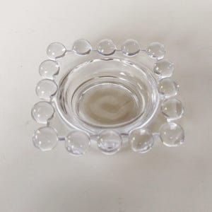 May include: Clear glass votive candle holder with a square shape and a beaded edge. The center is a circular well to hold a candle. The glass is transparent, allowing light to pass through.