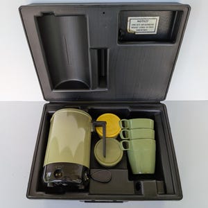 Puede incluir: Un juego de cafetera portátil vintage verde oliva y negro, que incluye una cafetera, tazas y tapas, cuidadosamente organizado dentro de un estuche de transporte negro. El estuche tiene un compartimento con una etiqueta de aviso.