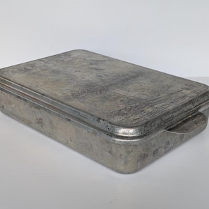 May include: A rectangular, weathered aluminum baking pan with a matching lid. The metal surface shows signs of age, including dark spots, scratches, and a dull, matte finish. It features two small, flat handles on the sides for easy lifting.