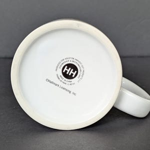 May include: White ceramic mug with a rounded handle, viewed from the bottom. The base of the mug features a circular logo with the letters "HH" inside. Text around the logo reads "PRODUCED FOR HOUSTON HARVEST GIFT PRODUCTS LLC" and "MADE IN CHINA".