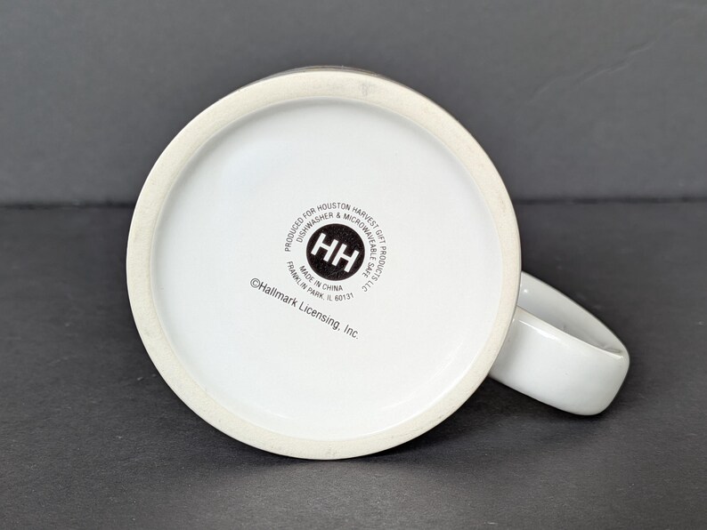May include: White ceramic mug with a rounded handle, viewed from the bottom. The base of the mug features a circular logo with the letters "HH" inside. Text around the logo reads "PRODUCED FOR HOUSTON HARVEST GIFT PRODUCTS LLC" and "MADE IN CHINA".