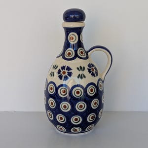 May include: A ceramic pitcher with a handle and a lid. The pitcher is primarily cream-colored with a dark blue pattern of circles and floral designs. The lid and top of the pitcher are dark blue.