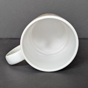 May include: A white ceramic mug with a rounded handle, viewed from above. The interior of the mug is white, and the handle is smooth and curved. The mug is set against a dark gray background.