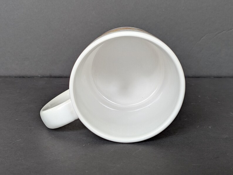 May include: A white ceramic mug with a rounded handle, viewed from above. The interior of the mug is white, and the handle is smooth and curved. The mug is set against a dark gray background.
