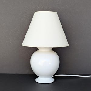 May include: A small, white table lamp with a rounded base and a conical lampshade. The lamp is made of glossy white material and has a simple, elegant design. The lamp is plugged in and ready to use.