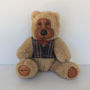 May include: A plush teddy bear with a wooden face and paws. The bear wears a plaid vest and a red bow tie. The bear's paws have a wooden design and the words "Robert Raikes" are visible on one paw.