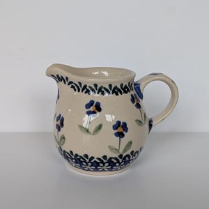 May include: A cream-colored ceramic creamer pitcher featuring a hand-painted pattern of small blue flowers with green leaves and yellow centers. The pitcher has a curved handle, a pouring spout, and a decorative blue floral border at the top and base.
