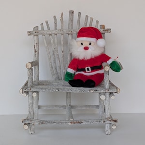 May include: A rustic, white-washed wooden chair with a Santa Claus plush toy. The Santa Claus toy is wearing a red suit, black belt, and green gloves. The chair is made of natural branches and has a unique, handcrafted appearance. A festive Christmas decoration.