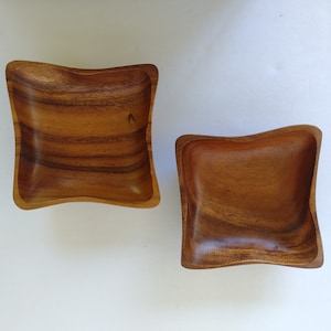 Pode incluir: Duas tigelas de madeira com uma forma ondulada, como uma almofada. As tigelas são de madeira marrom escura e estão vazias.