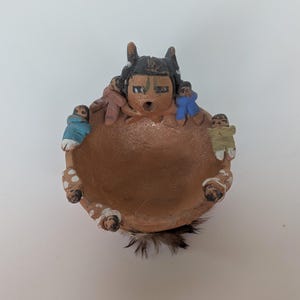 May include: A round, tan clay bowl featuring a central figure with a dark head and painted facial details. Several small, stylized figures in blue, tan, and white clothing are attached to the rim. Dark feathers are visible at the base of the bowl.