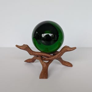 May include: A dark green glass sphere sits on a brown wooden stand. The sphere reflects the surrounding environment. The stand is made of intricately carved wood, with a natural, organic design. The sphere and stand create a decorative display.