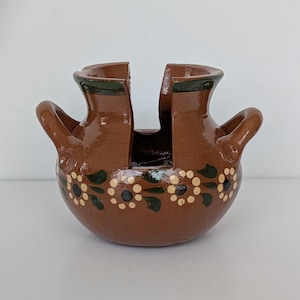 May include: A brown ceramic vase with two openings and two handles. The vase is decorated with a floral pattern in white, black, and green. The rim of the vase is painted green.
