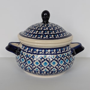 May include: A ceramic soup tureen with a lid, featuring a blue and white floral pattern. The tureen has two handles and a black knob on the lid. The design includes diamond and teardrop shapes. The tureen is likely used for serving soups or stews.