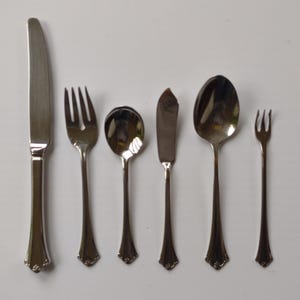 May include: A set of six silver-colored utensils, including a knife, two forks, two spoons, and a butter knife. The utensils have a decorative floral design at the base of the handles. The arrangement is on a white surface.