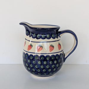 Op de afbeelding: Een keramische kan met een donkerblauwe rand en handvat. De kan heeft een crèmekleurige body met een patroon van rode aardbeien en donkerblauwe stippen. De kan heeft een schenktuit.