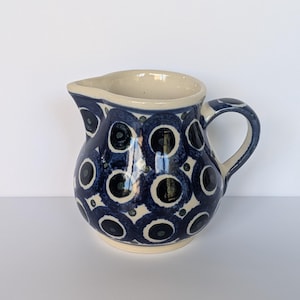 Może przedstawiać: Ceramiczny dzbanuszek na śmietankę o kremowej podstawie z powtarzającym się wzorem ciemnoniebieskich kół i małych kropek. Dzbanuszek ma zaokrąglony korpus, mały dzióbek do nalewania oraz zakrzywione ucho po prawej stronie.
