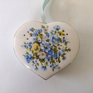 May include: A white ceramic heart-shaped box with a floral design. The box has blue and yellow flowers with green leaves. The box has holes around the edge.